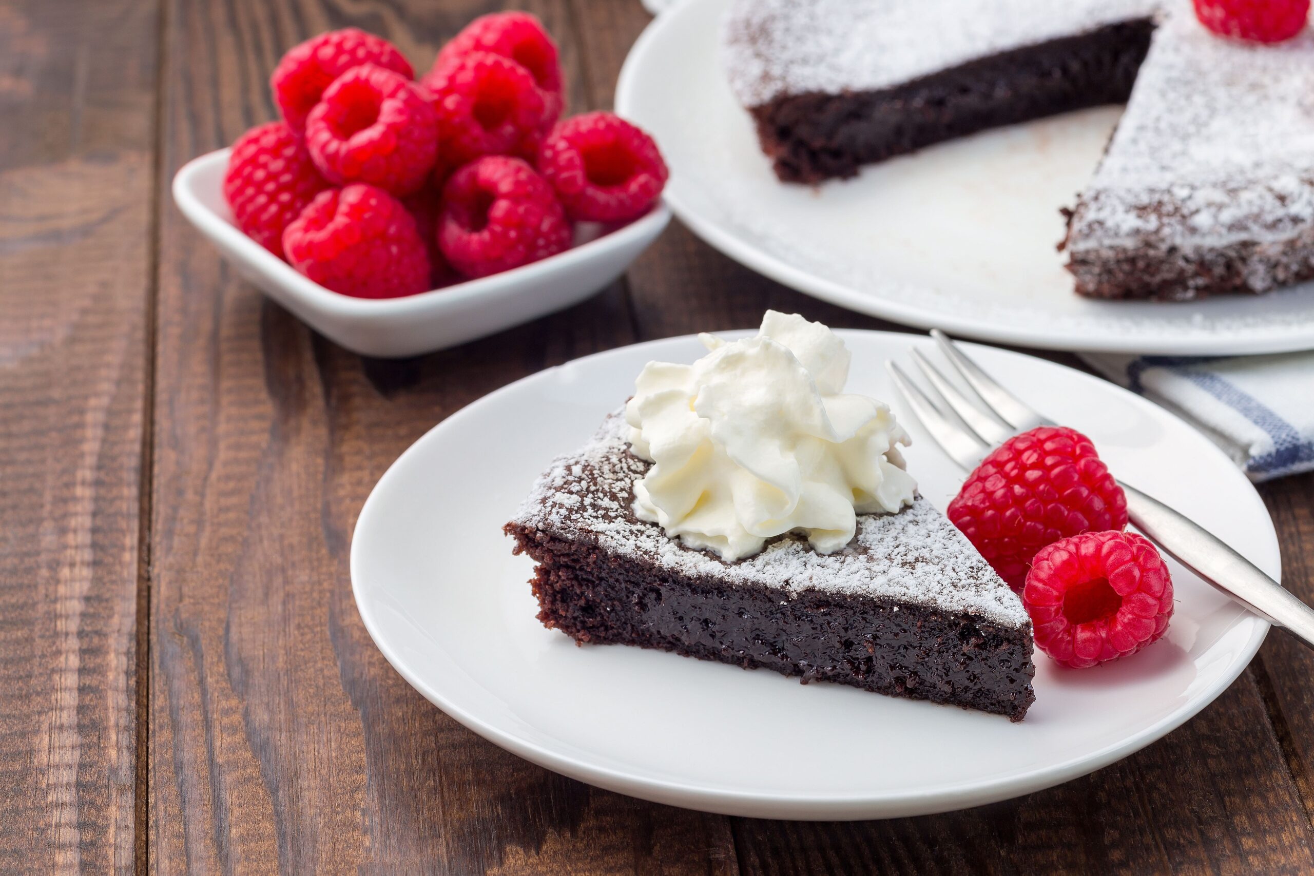 Piece of chocolate sticky brownie cake, swedish dessert Kladdkaka, on the plate, garnished with icing sugar, whipping cream and raspberry, horizontal